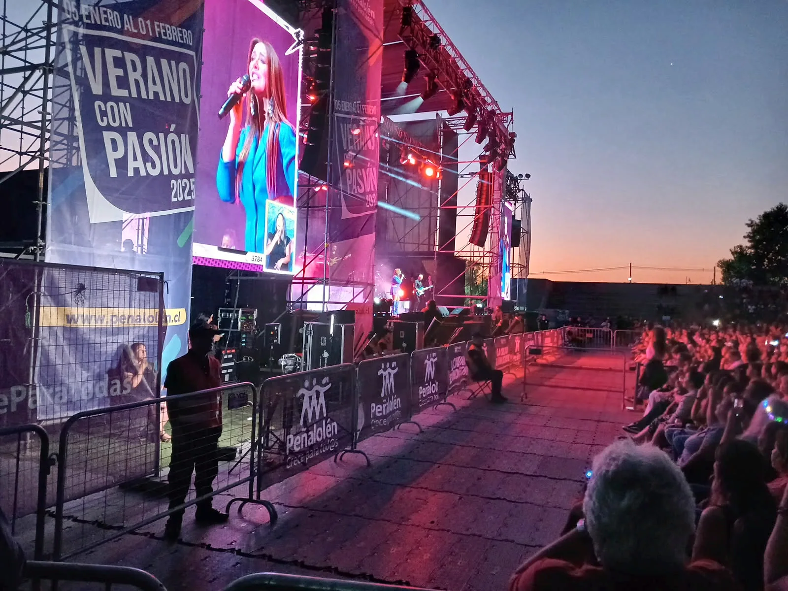 Escenario iluminado durante concierto al atardecer con vigilancia en lateral